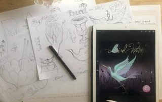 A workspace with sketches of various whimsical birds and plants on paper, a digital tablet displaying a colorful illustration of a bird, and drawing tools including a pen.