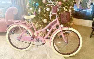 A pink bicycle with a brown seat and handles parked in front of a beautifully decorated christmas tree in a cozy living room setting, reminiscent of Melanie Stimmell's surrealism.