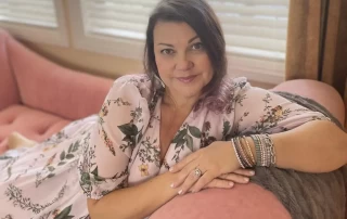 A woman with shoulder-length hair sits comfortably, smiling at the camera, leaning on a pink sofa arm. She wears a floral dress and multiple bracelets. Blinds partially filter the natural light in the background