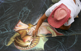 An artist in a pink hat draws a whimsical chalk portrait of a woman on an asphalt surface in Park West. The portrait features vivid colors and intricate details.