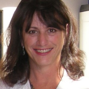 A middle-aged woman with shoulder-length brown hair and bangs, smiling at the camera. She wears a whimsical white top and earrings, with a silver necklace.