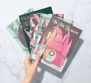 A hand holding a stack of three whimsical Christmas cards with colorful illustrations, including candy canes and holiday greetings, against a marble background.