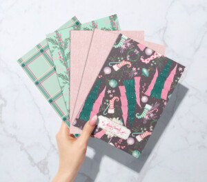 A hand holding a stack of festive Christmas-themed notebooks with patterns of candy canes, pine trees, and surrealist ornaments on a marble surface.