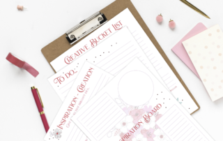 A neatly arranged desk with a clipboard holding a "creative bucket list" paper, surrounded by a pink notebook, gold pen, whimsical floral decorations, and office supplies on a white background.