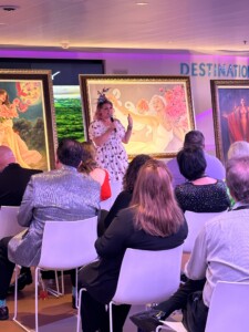 A woman in a polka dot dress and hat speaks to an audience at an art auction on a Park West cruise ship, with two framed surrealist paintings displayed behind her.