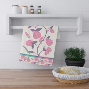 A stylish kitchen towel with a floral and fruit design by Melanie Stimmell hangs on a white wooden rack above a countertop. Beside it is a green potted plant and a basket with a