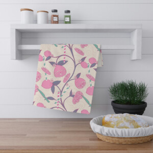 A colorful dishcloth with a vibrant pink and yellow fruit pattern hangs on a kitchen rack. Nearby, a surrealism-inspired potted green plant and a bread basket are placed on a wooden countertop against