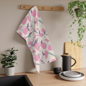 A decorative pink kitchen towel with a whimsical bird and fruit pattern, designed by Melanie Stimmell, hangs on a wooden rack in a modern kitchen, surrounded by houseplants and white dishes.