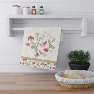 A floral embroidered towel hangs from a white shelf in a bright kitchen, beside it a potted plant and a surrealism-inspired basket of bread on the countertop.