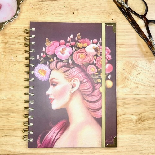 A spiral-bound notebook lies on a wooden surface. The cover features an illustrated profile of a woman with flowing pink hair adorned with flowers and butterflies. To the right, a pair of eyeglasses rests near the notebook.